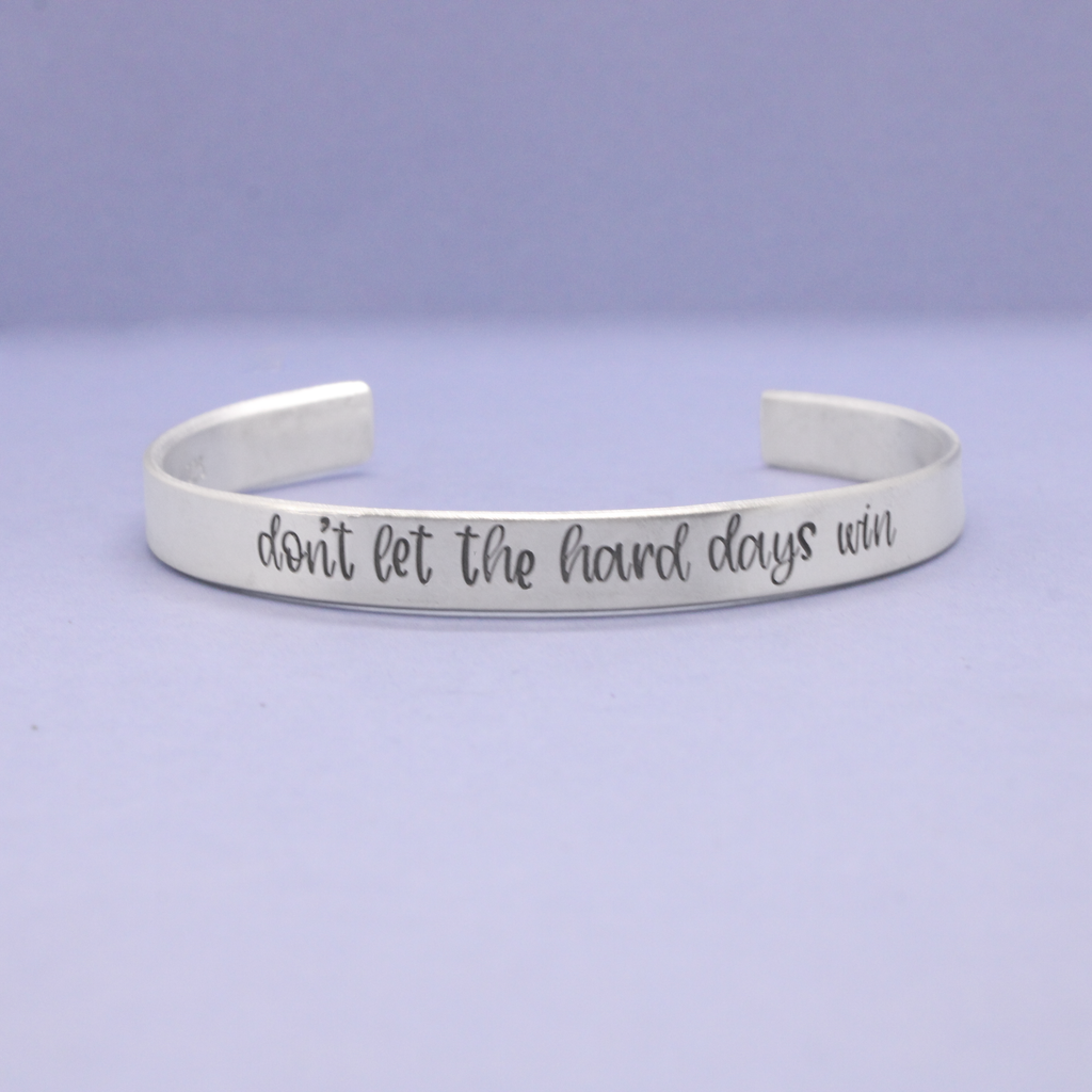 Silver bracelet with 'don't let the hard days win' text on a light purple background