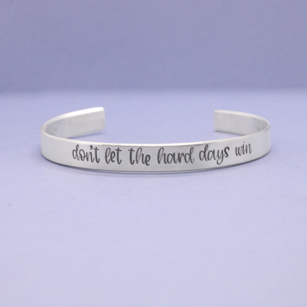 Silver bracelet with 'don't let the hard days win' text on a light purple background