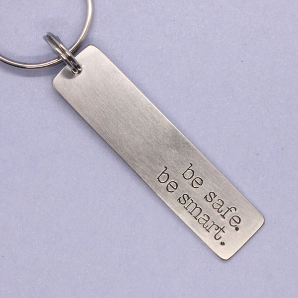 Silver keychain with 'be safe be smart' engraving on a light purple background