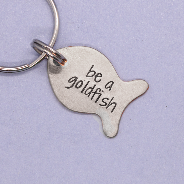 Silver fish-shaped keychain with 'be a goldfish' text on a light purple background