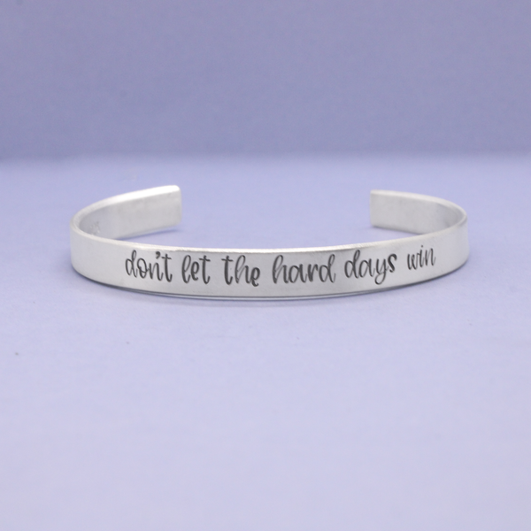 Silver bracelet with 'don't let the hard days win' text on a light purple background