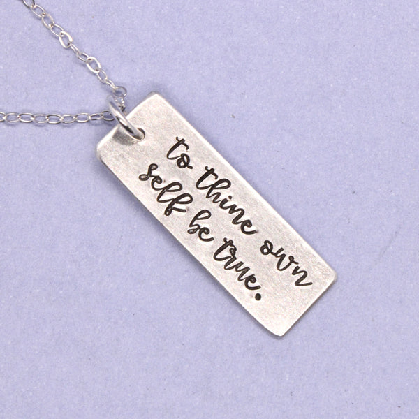 Silver necklace with a rectangular pendant engraved with 'to thine own self be true' on a light purple background.